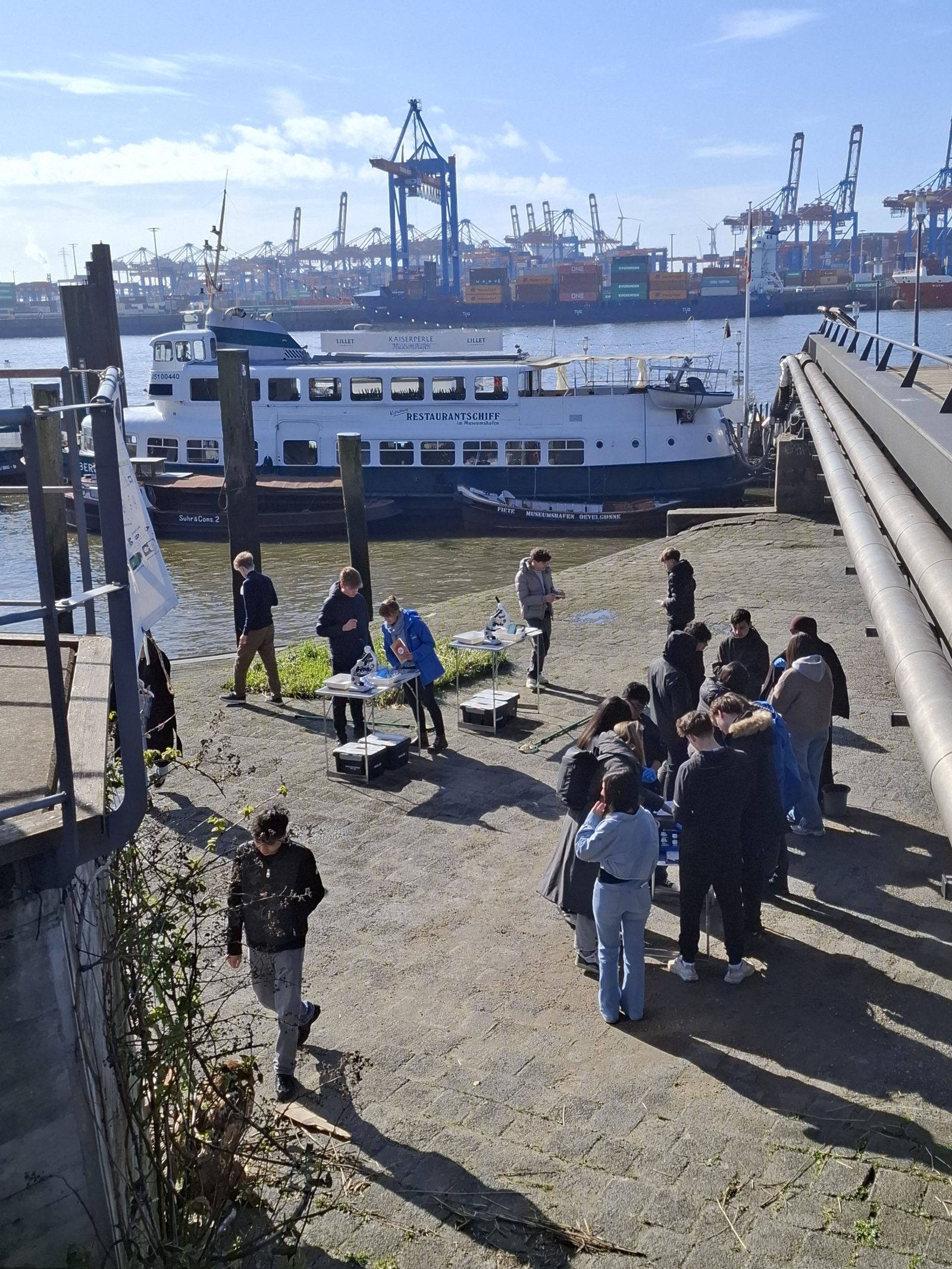 Gewässeruntersuchungen an der Elbe. Schüler_innen versammeln sich am Hamburger Hafen mit Containern im Hintergrund.
